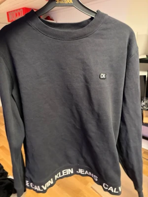 Svart sweatshirt från Calvin Klein - Svart långärmad sweatshirt från Calvin Klein med rund halsringning. Liten CK-logga på bröstet och Calvin Klein Jeans-tryck längs nederkanten. Mjuk bomullskänsla och klassisk passform, perfekt till jeans eller joggers.