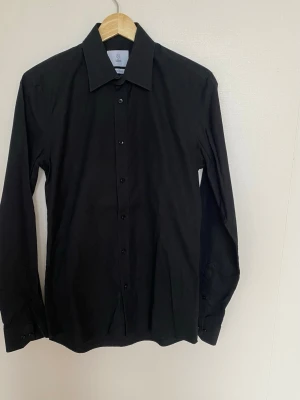 Svart skjorta från Bläck, Tailored Fit - Klassisk svart skjorta från Bläck med lång ärm och skräddarsydd passform. Skjortan har en stilren krage och knappar framtill.  Perfekt för dig som gillar en clean och enkel look.