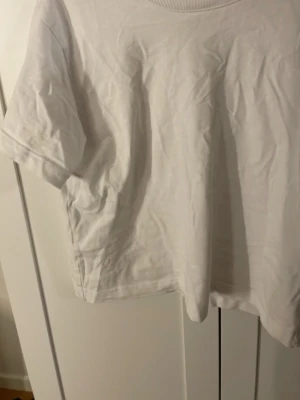 Vit basic t-shirt från Gina Tricot - En klassisk vit t-shirt från Gina Tricot, basically basics-kollektionen. T-shirten har rund halsringning och korta ärmar. Tillverkad i mjuk bomull för en skön och avslappnad look. Perfekt att matcha med jeans eller kjol.