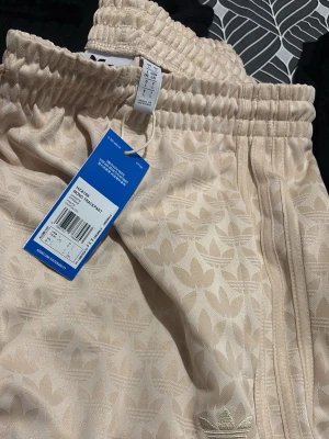 Beige shorts från Adidas Originals - Snygga beige shorts från Adidas Originals med diskret mönster av loggor och klassiska tre ränder längs sidorna. Resår i midjan och broderad logga framtill. Perfekta för en avslappnad stil.