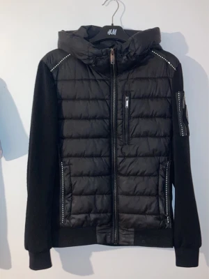 Svart vindjacka Supply & Demand - Svart vindjacka från Supply & Demand med vadderad front, huva och dragkedja. Jackan har detaljerade sömmar, fickor med dragkedja och en extra ficka på ärmen. Tillverkad i syntetmaterial och polyester för en sportig look.