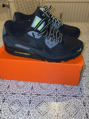 Nike Air Max 90 svart/grå sneakers - Nike Air Max 90 sneakers i svart och grått med reflekterande snören och neongula detaljer på tungan och framtill. Klassisk Air Max-sula med synlig luftbubbla och rund tå. Tillverkade i syntet och textil för en sportig look.
