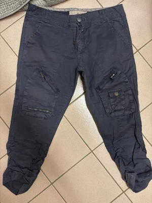 Mörkblå cargopants med dragkedjor - Snygga mörkblå cargopants med flera fickor, dragkedjor och knappdetaljer. Byxorna har en loose passform och croppad längd med justerbara benslut. Perfekta för en avslappnad och trendig stil.