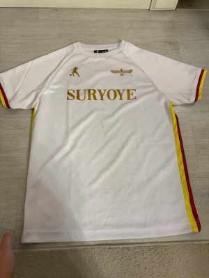 Vit Suryoye tröja - Vit sporttröja från LAIB med texten 'SURY0YE' i guld på bröstet, guldigt emblem och detaljer i rött och gult längs ärmarna. Aldrig använd (549kr nypris)