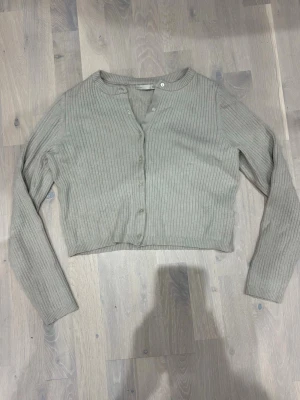 Grå ribbad kofta från 157 - Snygg grå ribbstickad kofta från 157 med lång ärm och croppad passform. Koftan har rund halsringning och knäppning framtill med små knappar. Perfekt att slänga över en topp för en chill look.