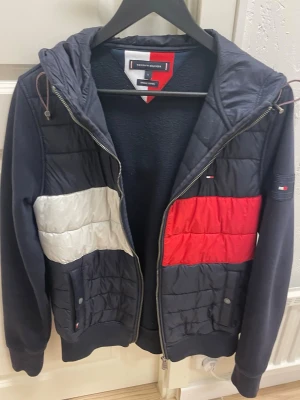 Tommy Hilfiger vindjacka med färgblock - Snygg vindjacka från Tommy Hilfiger med färgblock i rött, vitt och marinblått. Jackan har huva med snörning, dragkedja och två fickor framtill. Tillverkad i bomull och polyester.