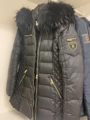 Svart dunjacka från Rockandblue - Svart dunjacka från Rockandblue med quiltad design, guldfärgade dragkedjor och stor pälskantad huva. Jackan har flera fickor med dragkedja och Rockandblue-logga på ärmen. Perfekt för kalla dagar.