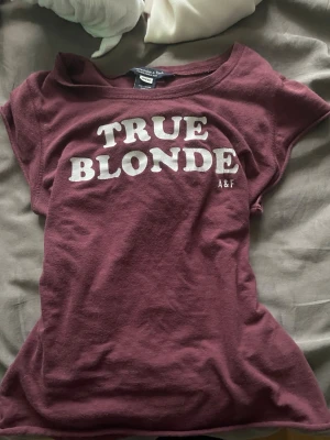 Vinröd t-shirt från Abercrombie & Fitch - Snygg vinröd t-shirt med vit text 'TRUE BLONDE' och A&F-logga på bröstet. Klassisk rund hals och korta ärmar. Perfekt för en avslappnad och trendig stil. Sitter mer som en S än M