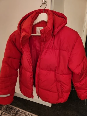 Röd pufferjacka från Lindex - Snygg röd pufferjacka från Lindex med huva och dragkedja framtill. Jackan har quiltad design och reflekterande detalj på ena ärmen. Perfekt för kalla dagar och ger en cool look.