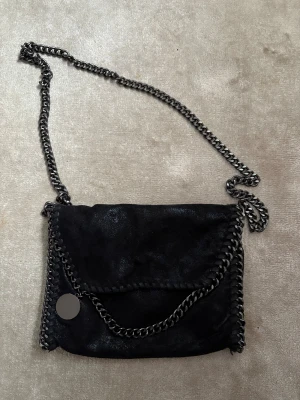 Svart axelväska med kedjedetaljer - Snygg svart axelväska med glansig mockaliknande yta och grova kedjor längs kanterna och som axelrem. Väskan har ett runt metallhänge och insidan är fodrad med leopardmönstrat tyg samt har en innerficka med dragkedja. Perfekt för dig som gillar coola detaljer.