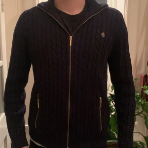 Mörkblå kabelstickad kofta Ralph Lauren - Kabelstickad mörkblå kofta från Ralph Lauren med guldfärgade dragkedjor och RL-broderi på bröstet. Tillverkad i 100% bomull, har långärmad design och ribbade muddar. Perfekt för dig som gillar klassisk stil med snygga detaljer.