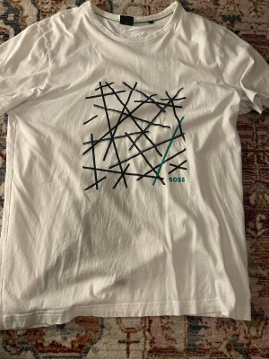 Vit t-shirt med grafiskt tryck från BOSS - Vit t-shirt från BOSS med coolt grafiskt tryck i svart och grönt på framsidan. Rund halsringning och korta ärmar. Materialet är mjuk bomull som känns skönt mot huden. Perfekt för dig som gillar stilrena plagg med en twist.