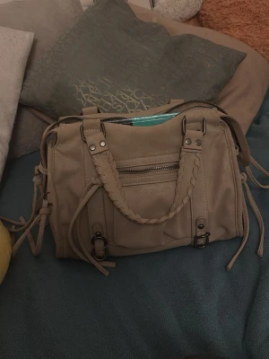 Beige axelväska med flätade detaljer - En jätte fin väska som får plats med mycket saker tillexempel skolböcker. Väskan är nästan helt ny och säljer då den inte kommer till användning. Skriv privat för fler bilder eller frågor!