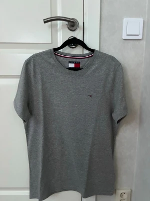 Grå t-shirt Tommy Jeans - Snygg grå t-shirt från Tommy Jeans med klassisk rund hals och diskret logga broderad på bröstet. T-shirten är kortärmad och har en stilren design med mjukt bomullsmaterial. Perfekt för dig som gillar enkel och tidlös stil.