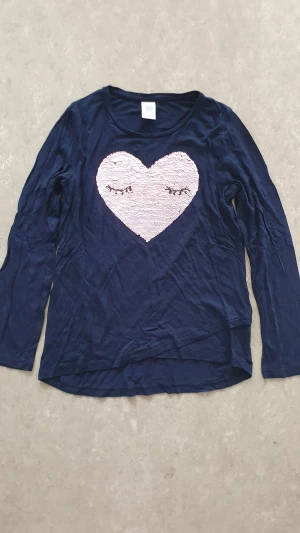 Tröja - Mörkblå långärmad tröja från Lindex med ett stort rosa hjärta i paljetter på bröstet. Hjärtat kan vändas och visar antingen sovande ögon eller texten HELLO. Tillverkad i mjuk bomull, perfekt för dig som gillar detaljer som sticker ut.