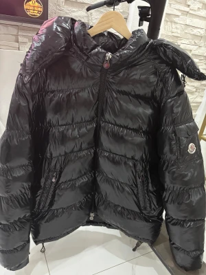 Svart Moncler pufferjacka med huva - Svart glansig pufferjacka från Moncler med huva och dragkedja framtill. Jackan har Moncler-logga på ärmen, sidofickor med dragkedja och quiltad design. Har använt den bara förra vintern några gånger o de va de. Så den är helt ny och säljer till biligt pris 700kr Fynd!!!! Har 2 st i storlek 2xl