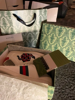 Gucci Ace sneakers  - Gucci Ace sneakers i vitt skinn med broderad röd och svart orm på sidan, klassiska gröna och röda ränder samt metallicröd och metallicgrön häl med reptilmönster. Platt sula, snörning och rund tå. Snygga detaljer och lyxig känsla, perfekt för dig som vill sticka ut. Pric kan diskuteras 