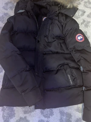 Svart dunjacka från Canada Goose - Säljer en svart dunjacka från Canada Goose med pälskant på huvan och klassisk logga på ärmen. Jackan har flera fickor, dragkedja och knappar framtill. Perfekt för kalla vinterdagar och har en riktigt schysst puffad look.