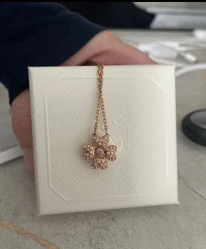 Swarovski Idyllia blomformad halsband - Snyggt halsband från Swarovski med ett blomformat hänge i guldton och glittrande rosa stenar. Kedjan är tunn och elegant med små detaljer och logotyp. Perfekt för dig som gillar blommor och bling.