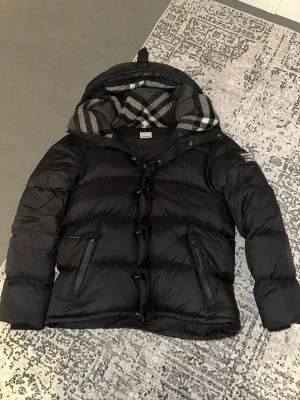 Svart Burberry dunjacka med avtagbara ärmar - Svart dunjacka från Burberry med avtagbara ärmar, så du kan bära den som väst. Jackan har huva med klassiskt rutigt Burberry-foder, dragkedja och knappar framtill samt två fickor med dragkedja. Perfekt för kalla dagar och har en riktigt schysst look.