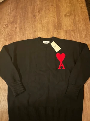 Svart stickad tröja från Ami - Svart stickad tröja från Ami Alexandre Mattiussi med rund hals och ribbade muddar. På bröstet finns ett stort rött hjärta med bokstaven A, vilket ger en unik och snygg detalj. Perfekt för dig som gillar stilrena plagg med en twist.