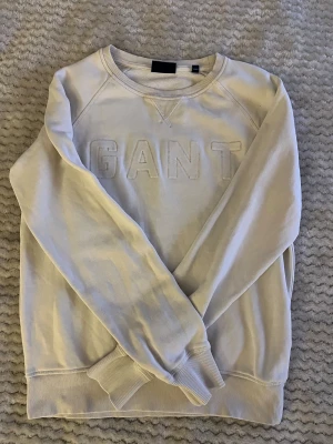 Vit sweatshirt från GANT - Säljer en stilren vit sweatshirt från GANT med broderad logga framtill. Tröjan har rund hals, ribbade muddar och långärmad passform. Perfekt för dig som gillar klassisk och enkel stil. Materialet är mjuk bomull som känns skönt mot huden.