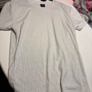 Vit t-shirt med diskret mönster Black Squad - Vit t-shirt från Black Squad. T-shirten har korta ärmar och är täckt av ett subtilt, upprepat RS-mönster som ger en cool textur. Rund halsringning och mjukt bomullsmaterial gör den skön att bära. Perfekt för dig som gillar stilrena detaljer.