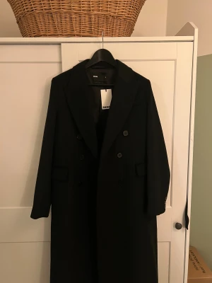 Hellång kappa i ull från Bik Bok, nyskick - Svart hellång ullkappa i klassisk och lyxig modell ”Phoenix” från Bik Bok i storlek S. Kappan har en rak och stilren passform. Helt oanvänd, nypris 1600.