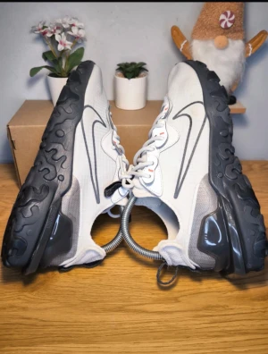 Nike React Element sneakers vita - Nike React Element sneakers i ljusgrått och vitt med svarta detaljer och sula. Snyggt mönstrad mesh-överdel, stora svarta swoosh-loggor på sidorna och coola röda accenter vid snörningen. Platt sula med unik struktur och dragflik bak. Perfekta för dig som gillar streetstyle.