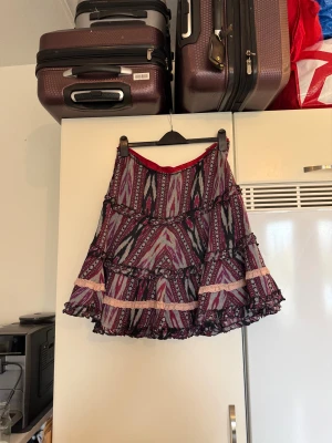 Mönstrad volangkjol med spetsdetaljer - Kort kjol med färgstarkt mönster i vinrött, svart och grått. Kjolen har flera volanger och ljusrosa spetsdetaljer som ger en cool och bohemisk vibe. Midjan är markerad med ett vinrött band och materialet känns lätt och luftigt, perfekt för dig som gillar att sticka ut. Från märket Odd Molly. Storlek 3 som enligt deras storleksguide motsvarar en L. Mycket fint skick, jag hittar inga synliga anmärkningar eller defekter. 