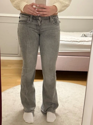Grå bootcut jeans från True Religion - Säljer ett par grå jeans från True Religion med bootcut passform och låg midja. Jeansen har klassiska fem fickor, snygga detaljer på bakfickorna och silverfärgade knappar. Materialet är jeans med en mjuk tvättad look. Tycker dessa jeans är skit snygga, säljer dom pågrund av att de har blivit för små på mig. 
