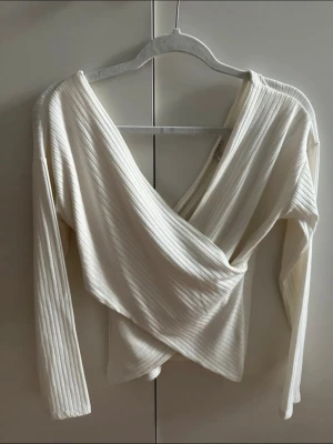 Vit omlottopp med ribbad struktur - Säljer en snygg vit omlottopp från Nelly Trend med ribbad struktur. Toppen har lång ärm och djup v-ringning som ger en cool och modern look. Perfekt att styla med byxor eller kjol