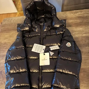 Svart pufferjacka från Moncler - Säljer en svart pufferjacka från Moncler med glansig finish och huva. Jackan har dragkedja framtill, ikonisk Moncler-logga på ärmen och coolt serietryck på insidan. Perfekt för dig som vill ha en varm och snygg jacka med street vibe.
