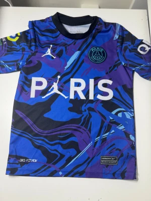 PSG Mbappé fotbollströja Nike 128 - Snygg PSG fotbollströja med Mbappé och nummer 7 på ryggen. Tröjan har ett blått, lila och svart abstrakt mönster, Paris Saint-Germain-logga och Nike Dri-Fit material som andas. Perfekt för dig som älskar fotboll och vill ha en cool look på planen.