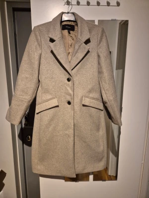Beige kappa från Vero Moda, M - Snygg beige kappa från Vero Moda i storlek M. Klassisk rak modell med bred krage, två svarta knappar framtill och fickor med lock. Materialet är mjukt och känns som ullblandning, perfekt för höst och vinter. Stilren och enkel att matcha med olika outfits.