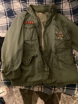 Grön parkajacka Polo Ralph Lauren - Säljer en grön parkajacka från Polo Ralph Lauren med coolt broderat motiv av en kanin och text på både fram- och baksidan. Jackan har flera stora fickor, snörning i midjan och dragkedja framtill. Perfekt för dig som gillar street och unika detaljer. Finns inte heller på marknaden längre. Köptes för 2700 men säljer för 2000, säljer enbart i Stockholm 