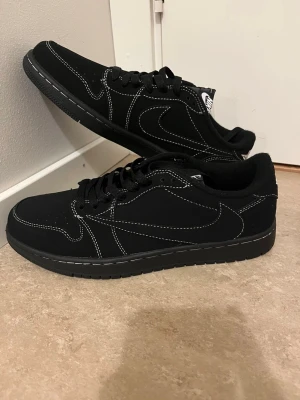 Nike Air Jordan 1 Low svart mocka - Svarta Nike Air Jordan 1 Low sneakers i mocka med vita kontrastsömmar och broderade detaljer på hälen. Rund tå, platt sula och klassisk snörning. Medföljer extra skosnören i rött och svart/vitt mönster. Snygg och stilren siluett som sticker ut.