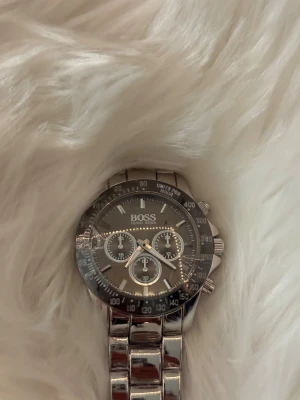 Hugo Boss klocka  - Stilren klocka från Hugo Boss i silverfärgat rostfritt stål. Klockan har svart urtavla med tre mindre visartavlor, tydliga index och tachymeter på ringen. Länkarmbandet är robust och glänsande, perfekt för dig som gillar en klassisk men modern look.