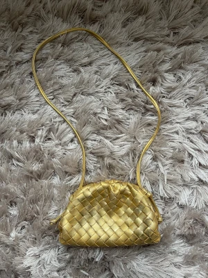 Guldig flätad väska  - Snygg axelväska i guldigt flätat skinn med lång rem. Väskan har en mjuk, rundad form och stängs med dragsko. Perfekt storlek för det viktigaste och ger en lyxig touch till din outfit.