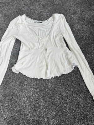 Vit croppad långärmad blus med spets - Säljer en söt vit blus från Gina Tricot med croppad passform och långa ärmar. Toppen har spetsdetaljer längs halsringningen och framtill, samt en liten rosett. Perfekt för dig som gillar romantisk och feminin stil.