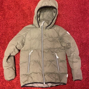 Olivgrön pufferjacka från Reima - Varm och go dunjacka från finska kvalitetsmärket Reima.    Avtagbar huva och dragkedja framtill. Jackan har två fickor med dragkedja och en innerficka. Reflexer på blixtlåsen och reflex på axel och rygg. Olivgrön.   Härligt varm. Vattenavvisande och vindtät.    Vadderingen är 60 % ankdun och 40 % andfjäder.