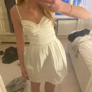 Vit kort klänning med smala axelband - Supersöt vit klänning med smala justerbara axelband, perfekt till studenten eller avslutning! Kjolen är kort och har en lätt puffig form med veck. 