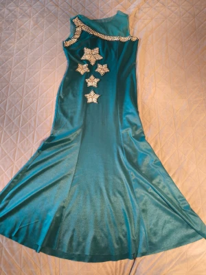 Turkos långklänning med stjärndetaljer - Snygg turkos långklänning i glansigt material med asymmetrisk axel och dekorativa stjärnor av paljetter framtill. Klänningen har en figurnära passform upptill och utsvängd kjol, perfekt för dig som vill sticka ut med coola detaljer.