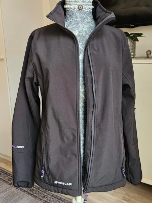 Svart vindjacka Whistler - Svart vindjacka från Whistler med fleecefoder, dragkedja framtill och på fickorna, samt snörning i nederkant. Jackan har hög krage och diskreta lila detaljer vid dragkedjorna. Perfekt för dig som vill ha en sportig och funktionell look.