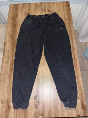 Mörkgrå mjukisbyxor från Gymshark S - Mörkgrå mjukisbyxor från Gymshark i storlek S. Byxorna har elastisk midja med snörning, muddar vid bensluten och två sidofickor. Diskret Gymshark-logga broderad på ena benet. Perfekta för chill eller träning.