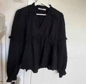 Svart volangblus från NA-KD - Supersnygg svart blus från NA-KD med volangdetaljer och v-ringning. Blusen har lång ärm med resår vid ärmslut och är gjord i ett lätt, krinklat tyg som ger en avslappnad men chic vibe. Perfekt att styla med jeans eller kjol för en trendig look. ( passar 34-36) eftersom den e lite större