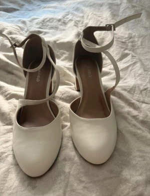 Vita pumps med klack från Anna Field - Snygga vita pumps från Anna Field med rund tå och bred klack. Skorna har stilrena remmar som korsar foten och spänns runt vristen. Tillverkade i syntetmaterial med en klassisk och elegant look. Perfekta för dig som vill ha en chic och bekväm sko med lite höjd.