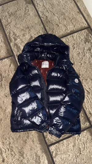 Moncler Maya  - Säljer en mörkblå pufferjacka från Moncler med glansig finish och huva. Jackan har dragkedja framtill, två sidofickor med dragkedja och ett brunt foder med Moncler-etikett och serietidningsdetalj. Perfekt för kalla dagar och riktigt snygg streetstil. Den är lite sönder men inget man tänker på, går att laga, ett litet jack bara