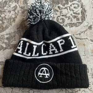 Svart och vit mössa med tofs från Allcap1 - Svart stickad mössa med vit text 'ALLCAP1' och broderad logga framtill. Mössan har en stor svartvit tofs på toppen och vita ränder runt om. Perfekt för kalla dagar och snygg streetstyle.