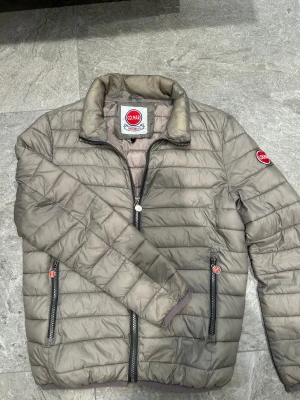 Jacka från Colmar - Snygg beige pufferjacka från Colmar med quiltad design och hög krage. Jackan har två dragkedjeförsedda fickor fram, Colmar-logga på ärmen och en innerficka. Materialet är lätt och perfekt för kyliga dagar. Passformen är normal och jackan har långa ärmar. Skick - 8/10
